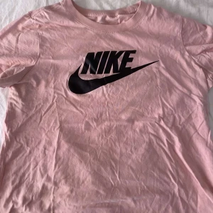 Rosa Nike t-shirt med logga - Säljer en rosa t-shirt från Nike med stor svart logga framtill. Klassisk rund halsringning och korta ärmar. Tillverkad i mjuk bomull som är skön mot huden. Perfekt för en avslappnad och sportig stil.