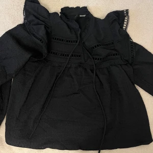Svart blus med volang och spetsdetaljer - Supersnygg svart blus från hm med volang vid axlarna, dekorativa spetsdetaljer och knytband vid halsen, köpt i Spanien för 31€ och säljs inte längre i svergie🩷aldrig andvänd, super snygg på och är i stolek s