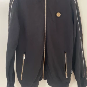 Svart Versace hoodie med gulddetaljer - Svart hoodie från Versace med guldfärgad Medusa-logga på bröstet och gulddetaljer längs ärmarna. Tröjan har hel dragkedja framtill, två fickor med dragkedja och ribbade muddar. Snygg och lyxig streetstyle med exklusiv känsla.