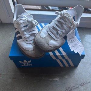 Adidas Samba vita sneakers - Klassiska Adidas Samba sneakers i vitt skinn med grå mockadetaljer och svarta ränder på sidorna. Låg modell med snörning, gummisula och rund tå. Ikonisk blå logga på plösen och guldfärgad 'SAMBA'-text på sidan. Perfekta för en clean och sportig stil.