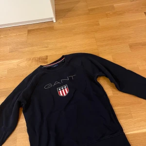 Mörkblå sweatshirt från GANT - Snygg mörkblå sweatshirt från GANT med klassiskt broderat logotyptryck och sköld på bröstet. Tröjan har rund halsringning, långa ärmar och ribbade muddar. Perfekt för en stilren och sportig look.