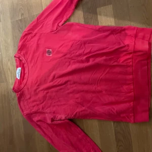 Röd Stone Island sweatshirt M - Röd sweatshirt från Stone Island med klassisk logotyp-patch på bröstet. Tröjan har rund halsringning och långa ärmar. Tillverkad i mjuk bomull som är skön att bära. Perfekt för dig som gillar streetwear och vill sticka ut med en färgstark look.