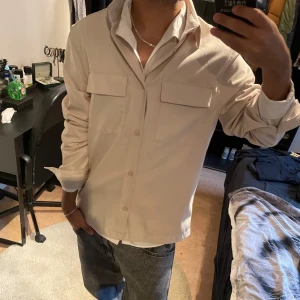 Beige overshirt med fickor  - En helt ny, Snygg beige overshirt med klassisk krage och två stora bröstfickor med lock. Skjortan har knäppning framtill och långa ärmar. Perfekt att bära öppen över en t-shirt för en avslappnad stil.