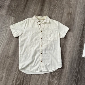 Vit kortärmad skjorta från Kappahl - Snygg vit skjorta från Kappahl i storlek 170. Skjortan har korta ärmar, klassisk krage, bröstficka och knappar framtill. Perfekt för en clean och stilren look under varmare dagar.