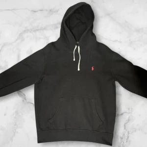 Svart hoodie från Ralph Lauren - Svart hoodie från Ralph Lauren med klassisk röd logga på bröstet och vit snörning i huvan. Tröjan har en stor magficka och långa ärmar, perfekt för chill dagar. Mjuk bomullskvalitet och normal passform. Nästan helt ny, använd cirka 10 gånger, kan tänka mig gå ner lite i pris för en snabb affär 