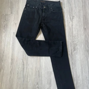 Svarta jeans från Nudie Jeans Co W31 L34 - Säljer ett par svarta jeans från Nudie Jeans Co i klassisk femficksmodell. Jeansen slim fit passform och är tillverkade i slitstark bomull. Snygga detaljer med Nudie-logga på bakfickan och läderpatch i midjan. Perfekt för dig som gillar stilrena och tidlösa jeans.