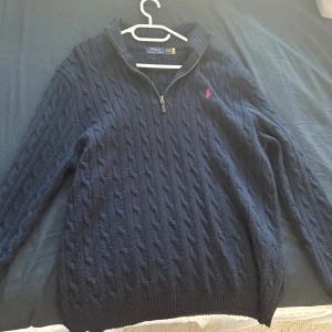 Halfzip Polo Ralph Lauren - Marinblå stickad tröja från Polo Ralph Lauren med klassiskt kabelstickat mönster och röd broderad logga på bröstet. Tröjan har lång ärm, ribbad krage och half zip-dragkedja framtill. Pris ej ristat i sten