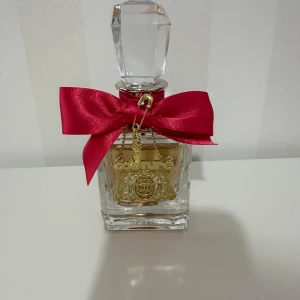 Juicy Couture Viva La Juicy parfym 50ml - Juicy Couture Viva La Juicy Eau de Parfum 50ml i en lyxig glasflaska med en stor, slipad kristallkork. Flaskan är dekorerad med en stark rosa rosett och guldfärgade hängen med logga och detaljer. Perfekt för dig som gillar söta och fruktiga dofter.