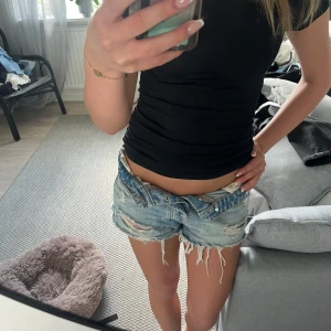 Blå slitna jeansshorts med fransar - Säljer ett par blå jeansshorts med låg midja och råa, fransiga kanter. Shortsen har slitningar och hål på framsidan för en riktigt chill och edgy vibe. Klassisk femficksmodell med knapp och dragkedja framtill. Perfekta för varma dagar och festivalhäng.