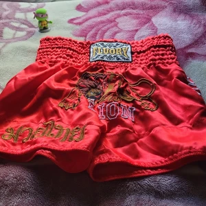 Röda Muay Thai shorts från Fluory - Snygga röda Muay Thai shorts från Fluory med broderad lejon-design och guldig text framtill. De har bred resår i midjan för extra komfort och är tillverkade i glansigt satinmaterial. Perfekta för dig som vill sticka ut på träningen!