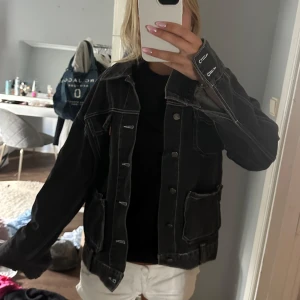 Svart oversized jeansjacka - Säljer en svart jeansjacka med kontrasterande vita sömmar och stora fickor framtill. Jackan har klassisk krage, knappar hela vägen och en oversized passform som ger en chill vibe. Perfekt att slänga över en hoodie eller t-shirt för en avslappnad look.