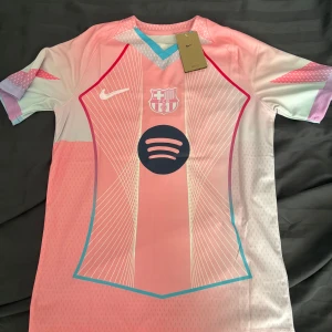 FC Barcelona Lamine Yamal rosa tröja Nike - Säljer en FC Barcelona fotbollströja från Nike med Lamine Yamal och nummer 10 på ryggen. Tröjan är rosa med vita och blå detaljer, tunna linjer och klubbmärke samt Spotify-logga på bröstet. Tillverkad i 100% polyester och har korta ärmar.