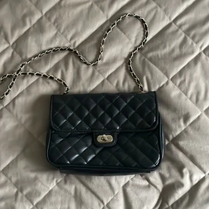 Svart quiltad axelväska med kedja - Säljer en svart quiltad axelväska med snygg kedjerem i guld och svart. Klassisk rektangulär form med lock och vridspänne i metall. Väskan har en lyxig känsla och passar perfekt till din outfit när du vill sticka ut lite extra.