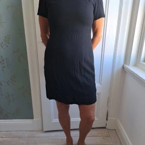 Svart kort klänning Mary Kay - En svart, enkel och klassisk kort klänning från Mary Kay by Brookhurst. Klänningen har korta ärmar, rund halsringning och rak passform. Tillverkad i ett mjukt tyg som ger en stilren look. Perfekt för dig som gillar minimalistisk stil.