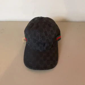Svart keps från Gucci med GG-mönster - Snygg svart keps från Gucci med klassiskt GG-monogram över hela kepsen. Kepsen har justerbart spänne bak och ikoniska gröna och röda ränder på sidan. Tillverkad i bomull och polyester, perfekt för dig som vill sticka ut med en lyxig accessoar.