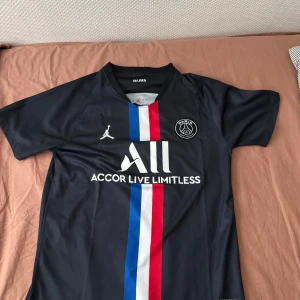 PSG x Jordan svart fotbollströja - Svart PSG fotbollströja från Jordan med vit, blå och röd rand längs mitten. Snyggt Paris Saint-Germain-emblem på bröstet och Jordan-logga. Tillverkad i lätt och ventilerande polyester, perfekt för match eller träning. Modern passform och korta ärmar.