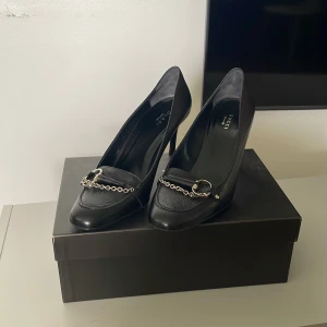 GUCCI klackar  - Eleganta och stiliga Gucci klackskor i helsvart. Skorna har en silvrig kjedja som detalj på framsidan och har bara andvänts vid få tillfällen. Skorna kommer även med den originals boxen och påsen till!