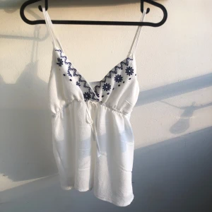 Vit linnetopp  - Superfin vit linnetopp med smala axelband som går att justera där bak och blå broderade blommor vid v-ringningen. Toppen har knyt under bysten.