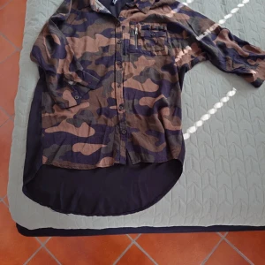 Camouflage skjorta FB Sister S - Cool lång skjorta från FB Sister i storlek S med brunt och grönt camouflage-mönster. Baksidan har ett stort tryck med texten 'Grunge Couture'. Skjortan har knappar framtill, bröstficka med dragkedja och lång bakdel. Materialet är lätt och känns som bomull.