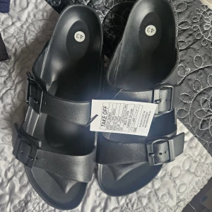 Svarta sandaler med dubbla spännen - Svarta sandaler från Take Off med två breda remmar och justerbara spännen. Tillverkade i syntetmaterial med platt sula och öppen tå, perfekta för varma dagar. Enkel och stilren design som passar till det mesta.