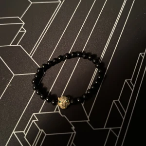 Svart pärlarmband med leopardhuvud - Coolt armband med svarta pärlor och en guldig leopardhuvuddetalj med gröna ögon. Perfekt accessoar för dig som vill sticka ut med en unik och djurinspirerad stil. Elastiskt band gör det enkelt att ta på och av.