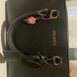 Svart handväska från Don Donna - Snygg svart handväska från Don Donna med guldfärgade detaljer . Väskan har två handtag, rymligt fack med dragkedja och flera innerfickor. Stilren design i skinnimitation som passar perfekt till din look.