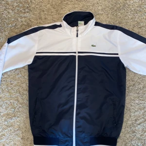 Blåvit vindjacka från Lacoste Sport - Snygg vindjacka från Lacoste Sport i marinblått och vitt med klassisk krokodillogga på bröstet och broderad Lacoste-text på ryggen. Jackan har hel dragkedja, ribbade muddar och sportig design med kontrastpaneler. 