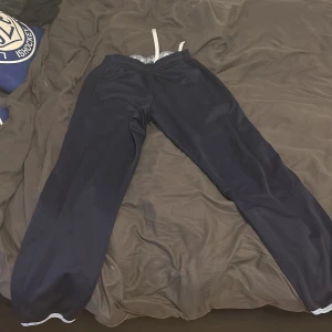 Svarta träningsbyxor från CCM - Svarta träningsbyxor från CCM med resår och vit dragsko i midjan. Byxorna har raka ben med resår vid bensluten och är tillverkade i ett syntetmaterial som passar perfekt för sport eller uppvärmning. 