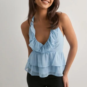 Ljusblå volangtopp från Nelly XS - Supersöt ljusblå topp från Nelly med volanger och djup v-ringning. Toppen har knyt i nacken, smala axelband och flera lager volang för en flowy look. Perfekt för dig som vill sticka ut med en feminin och trendig stil.