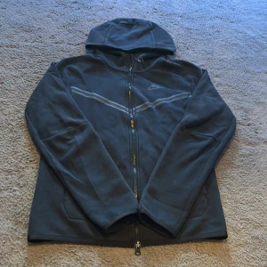 Svart fleecejacka med huva från Nike - Svart fleecejacka från Nike i storlek XS. Jackan har huva, hel dragkedja framtill och diskret Nike-logga på bröstet. Snygg detalj med reflekterande ränder över bröstet och en extra ficka med dragkedja på ärmen. Perfekt för kyliga dagar.