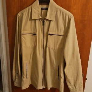 Beige overshirt från Dressmann M - Stilren beige overshirt från Dressmann i bomull, med dragkedja framtill och två bröstfickor med zip. Klassisk krage och långa ärmar med knapp vid ärmslut. Perfekt för lager-på-lager och enkel att matcha med jeans eller chinos.