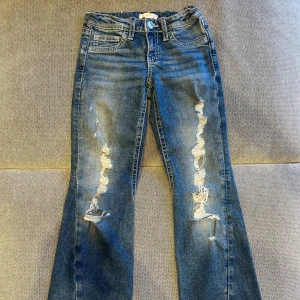 Blå bootcut jeans Gina Tricot 152 - Snygga blå bootcut jeans från Gina Tricot med slitna detaljer framtill och coola bakfickor med knappar. Jeansen har kontrastsömmar och klassisk femficksdesign. Perfekta för dig som gillar en avslappnad och trendig look.