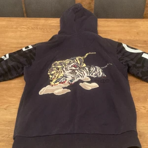 Svart hoodie med tigerbroderi från Kenzo - Säljer en svart hoodie från Kenzo med broderad tiger på ryggen och tigerhuvud på bröstet. Ärmarna har mörkt zebramönster och vita tryck. Hoodien har snörning och stor ficka fram. Perfekt för dig som gillar coola detaljer och streetwear.