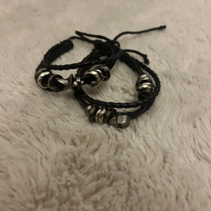 Svarta läderarmband med dödskalle & stjärna - Två svarta armband i flätat och slätt läder med coola detaljer i silverfärgad metall. Ena armbandet har en stjärna och det andra en dödskalle, båda med flera ringar som dekoration. Justerbara band för perfekt passform, perfekt för dig som gillar edgy stil.