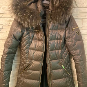 Rockandblue brun dunjacka med äkta päls - Snygg brun dunjacka från Rockandblue med guldig dragkedja, flera fickor och stor huva med fluffig fuskpäls. Jackan är quiltad och har en figurnära passform som ger en clean look. Perfekt för kalla dagar när du vill vara både varm och stilren.