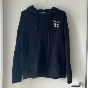 Svart hoodie House of Holland S - Svart hoodie från House of Holland med vit text 'Delete Your Idols' på bröstet och ryggen. Klassisk känguruficka, huva med snörning och långa ärmar. Perfekt för dig som gillar statement-plagg och streetstyle.