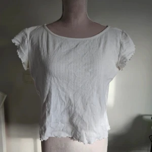 Vit pointelle topp från 157 L - Supersöt vit topp från 157 med pointelle-stickat mönster och korta ärmar. Den har en lätt croppad passform, vågig kant nertill och diskreta spetsdetaljer vid ärmslut. Perfekt för en fräsch och enkel look.