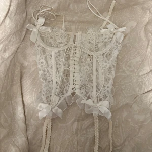 Vit spetskorsett från Victoria's Secret - Superfin vit korsett från Victoria's Secret med spets, snörning framtill och dekorativa rosetter. Modellen har justerbara axelband, volangkant och detaljer i satin. Perfekt för dig som vill ha en romantisk och feminin look.