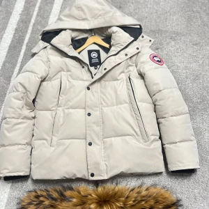Beige dunjacka från Canada Goose S - Säljer en beige dunjacka från Canada Goose med avtagbar huva och pälskant. Jackan har klassisk patch på ärmen, stora fickor och dragkedja med knappar. Perfekt för kalla vinterdagar och riktigt snygg till streetwear-stilen.