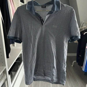Grå pikétröja från Hugo Boss - Snygg grå pikétröja från Hugo Boss med klassisk krage och diskret logga broderad på bröstet. Tröjan har korta ärmar, knappar vid halsen och är tillverkad i mjuk bomull. Perfekt för en clean och stilren look.