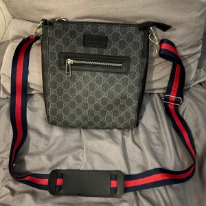  axelväska med GG-loggor - Snygg axelväska i svart med mönster och bred, justerbar axelrem i marinblått och rött. Väskan har dragkedja upptill, ytterfack med dragkedja och lyxigt fodrad insida med logga. Perfekt för dig som vill ha en stilren och ikonisk väska.