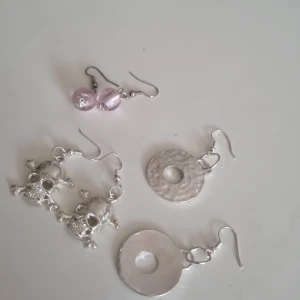 Tre par coola örhängen i silver och rosa - Säljer tre par örhängen: ett par med stora silverfärgade dödskallar, ett par med runda hamrade silverringar och ett par med rosa pärlor. Alla har krokar i metall och unika detaljer som sticker ut. Perfekt för dig som vill ha något annorlunda och edgy i smyckeslådan.
