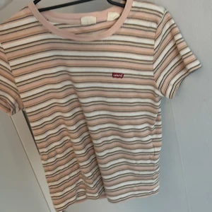 Randig t-shirt från Levi's - Säljer en snygg randig t-shirt från Levi's med korta ärmar. T-shirten har beige, vita och rosa ränder samt en liten röd Levi's-logga på bröstet. Mjuk och skön bomullskvalitet, perfekt till jeans eller shorts.