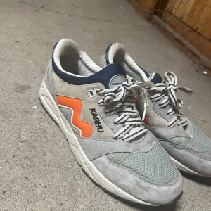 Karhu Aria 95 sneakers grå och orange - Karhu Aria 95 sneakers i grå mocka och mesh med orange detaljer på sidan och mörkblå inslag runt kragen. Skorna har snörning med vita och blå randiga skosnören, rund tå och platt sula. Perfekt för dig som gillar retrostil och snygga färgkombinationer.