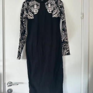 Svart klänning med leopardmönster ASOS - Cool svart långklänning från ASOS med långa ärmar och iögonfallande leopardmönster över axlar och ärmar. Framsidan har två stora leopardansikten som ger en edgy vibe. Perfekt för dig som vill sticka ut med en unik look.