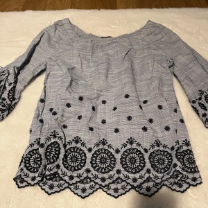 Blus med broderi och knytärmar - Säljer en ljusgrå blus med svarta broderade detaljer och blommönster. Blusen har offshoulder-modell, vida ärmar med knyt och vågig kant nertill. Tunt och luftigt material, perfekt för vår och sommar. Supersnygg till jeans eller kjol!
