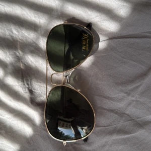 Guldiga aviator solglasögon - Snygga aviator solglasögon med tunna guldfärgade metallbågar och mörkgröna glas. Klassisk pilotform med svarta skalmar och näskuddar för extra komfort. Perfekta för att lyfta din stil i solen.