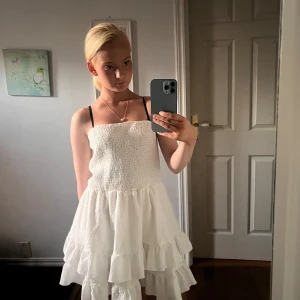 Vit volangklänning med smock - Supersöt vit klänning med smock upptill och volanger i kjolen. Axelbandslös design som ger en fräsch och somrig vibe. Perfekt för soliga dagar och chill häng. Materialet känns lätt och luftigt, och klänningen har en kort längd som gör den extra skön.