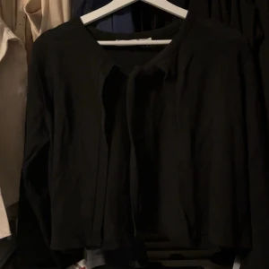 Svart kofta med knyt från 157 - Svart kofta från 157 i storlek S. Den har ett enkelt och stilrent utseende med knyt framtill vid halsen. Koftan är långärmad och gjord i ett mjukt stickat material som passar perfekt när du vill ha något bekvämt och snyggt.