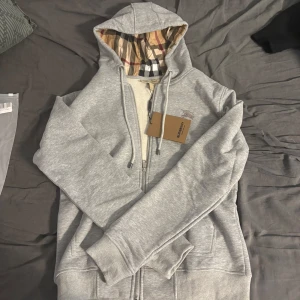 Grå hoodie från Burberry med dragkedja - Snygg grå hoodie från Burberry med klassiskt rutigt foder i huvan, broderad logga på bröstet och dragkedja framtill. Tröjan har snörning i huvan och ribbade muddar. Perfekt för dig som vill ha en stilren och exklusiv look.